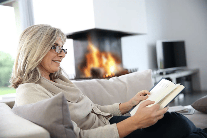 Senior woman reading book at home by fireplace copia